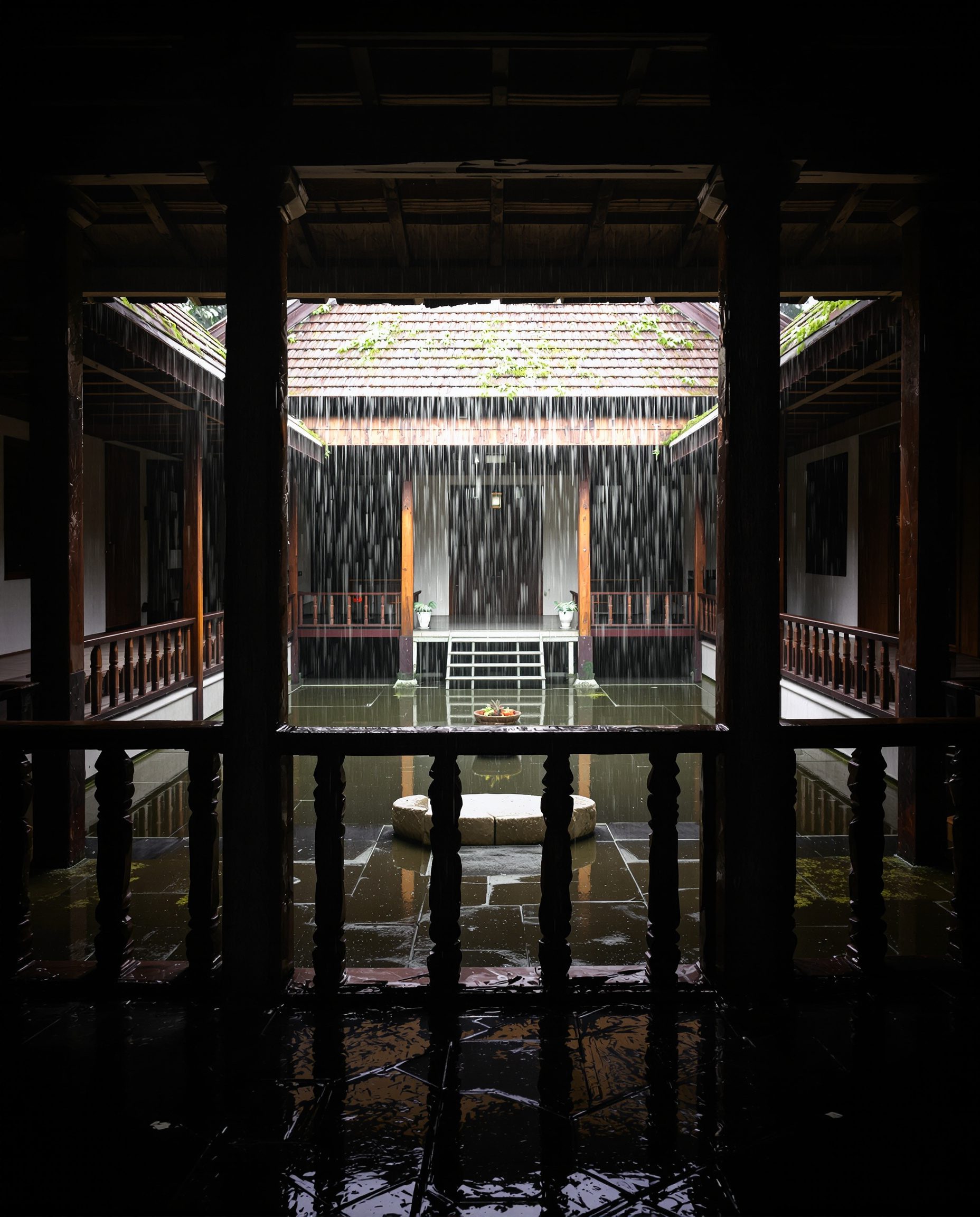 Rain falling into the open central courtyard of a traditional Kerala nalukettu, seen from the shaded verandah